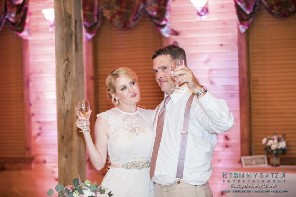 JenniferRudiWeddingReceptionPhotos-336 - TOMMY GATZ ENTERTAINMENT A bride and groom in a decorated venue raise wine glasses. The bride wears a sleeveless dress, and the groom is in a shirt with suspenders. There are floral arrangements in the background.