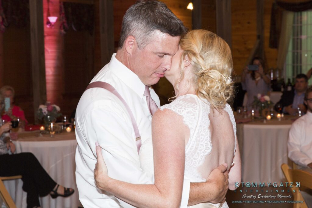 JenniferRudiWeddingReceptionPhotos-342 - TOMMY GATZ ENTERTAINMENT A couple embracing while dancing at an indoor wedding reception, with guests seated at tables in the background.