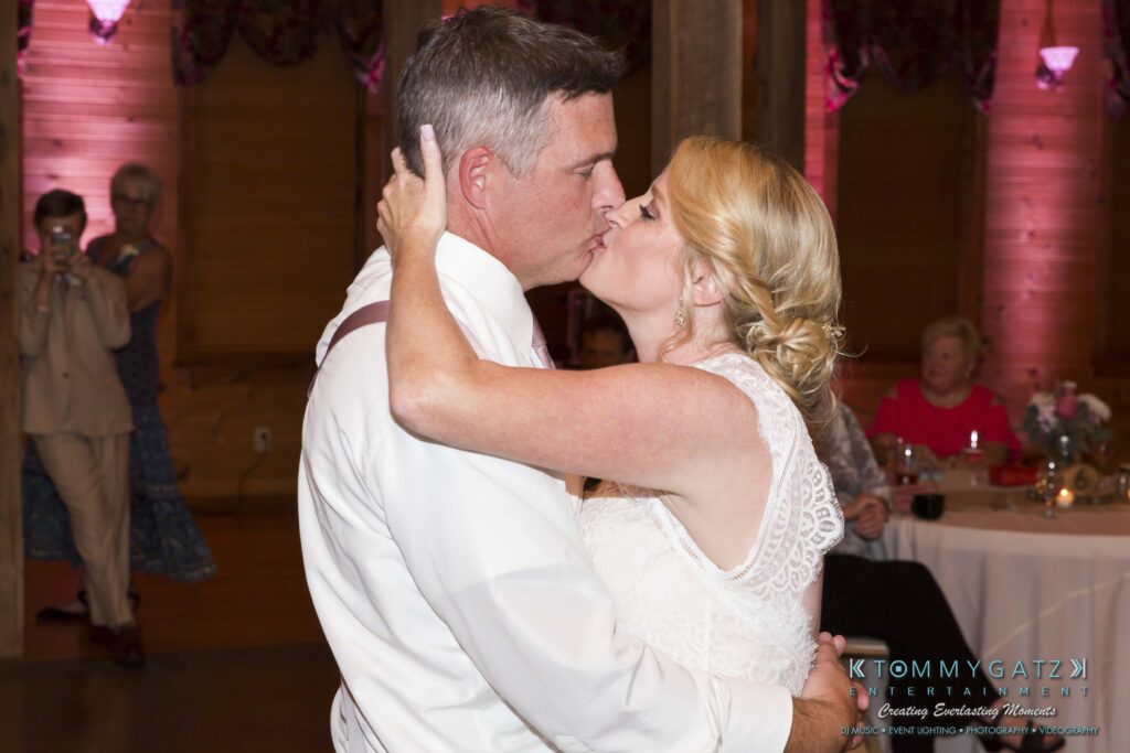 JenniferRudiWeddingReceptionPhotos-344 - TOMMY GATZ ENTERTAINMENT A couple shares a kiss while dancing at a wedding reception, with guests seated and another person taking a photo in the background.