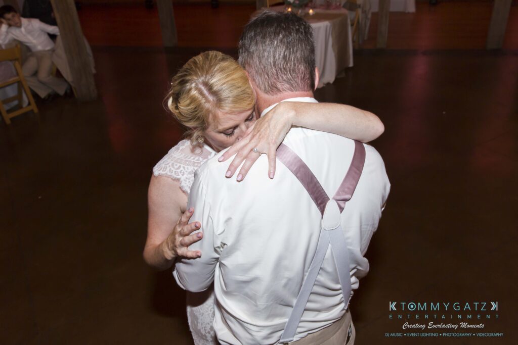 JenniferRudiWeddingReceptionPhotos-346 - TOMMY GATZ ENTERTAINMENT A couple embraces while dancing, with the woman resting her head on the man's shoulder. They are in a warmly lit room with wooden pillars.