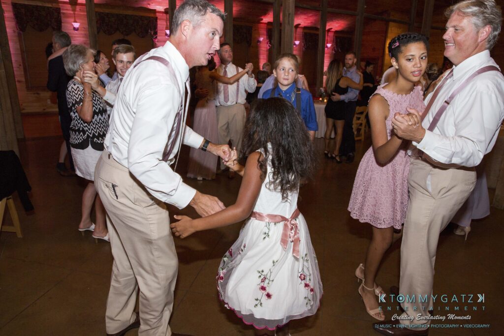 JenniferRudiWeddingReceptionPhotos-350 - TOMMY GATZ ENTERTAINMENT People dancing at an indoor event, wearing formal attire. Two men in white shirts hold hands with young girls in dresses. Other attendees converse and move around in the background.