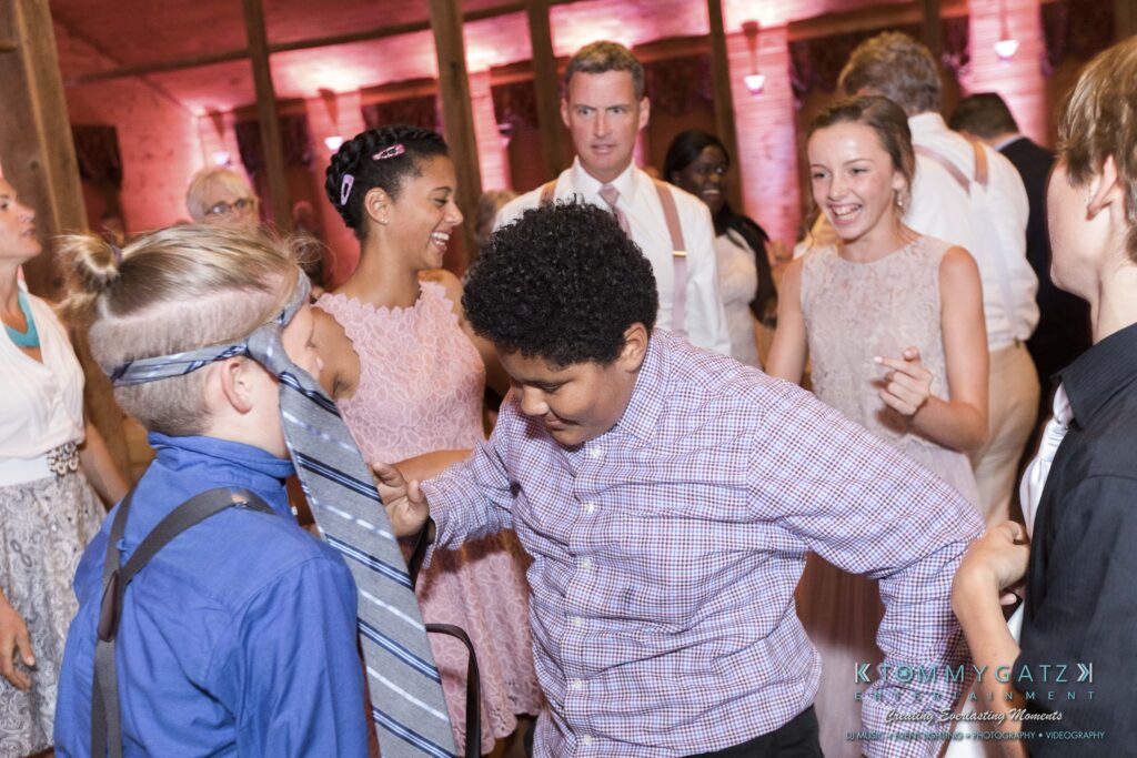 JenniferRudiWeddingReceptionPhotos-352 - TOMMY GATZ ENTERTAINMENT People dancing at an event, with one person wearing a tie over their eyes. The room has warm lighting and guests are dressed in formal attire.