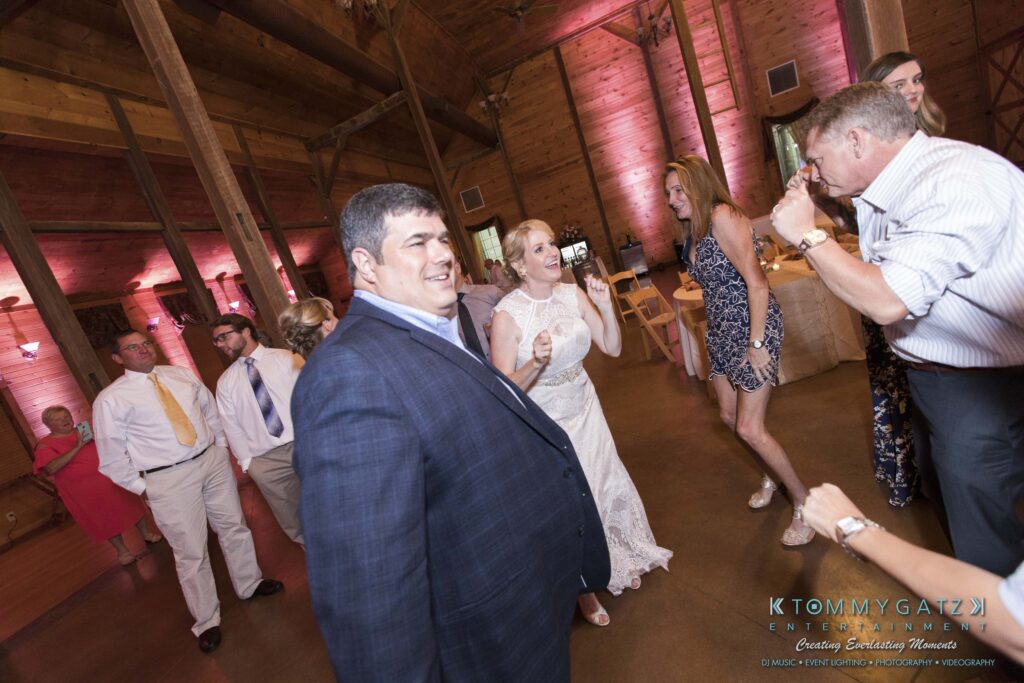 JenniferRudiWeddingReceptionPhotos-353 - TOMMY GATZ ENTERTAINMENT Wedding guests dancing energetically in a rustic venue with wooden beams and pink lighting. People are smiling and wearing formal attire.