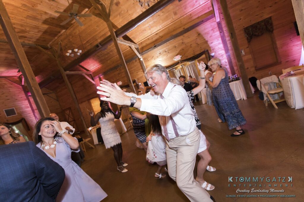JenniferRudiWeddingReceptionPhotos-355 - TOMMY GATZ ENTERTAINMENT A group of people dancing energetically in a wooden venue with a high ceiling. A man in a white shirt is prominent in the foreground, smiling with arms raised.