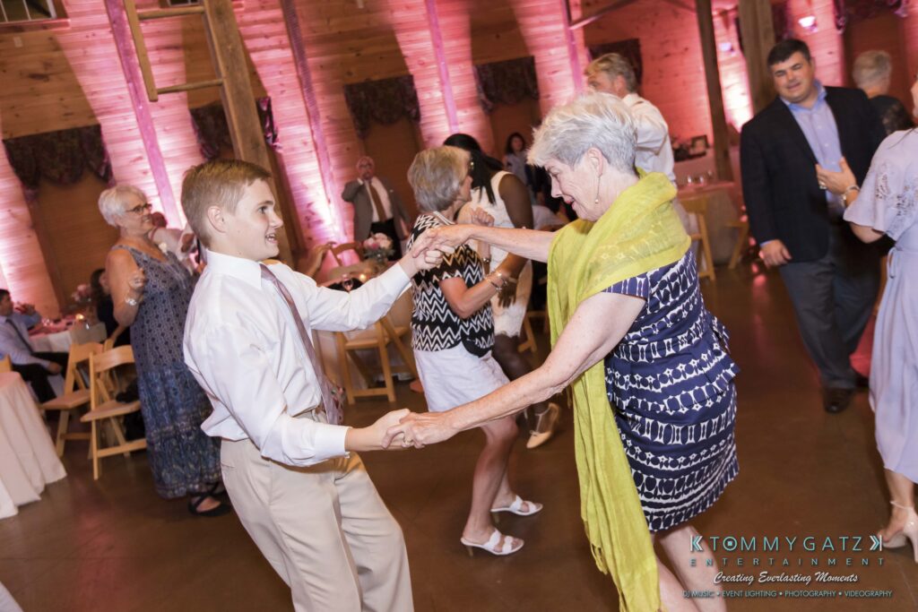 JenniferRudiWeddingReceptionPhotos-357 - TOMMY GATZ ENTERTAINMENT A young man and an older woman dance enthusiastically together in a warmly lit event space, surrounded by other guests.