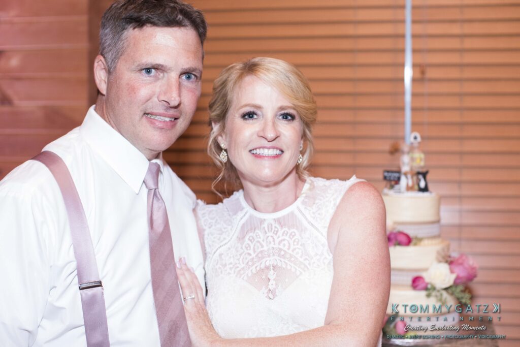 JenniferRudiWeddingReceptionPhotos-367 - TOMMY GATZ ENTERTAINMENT A couple poses together in formal attire with a tiered cake in the background.