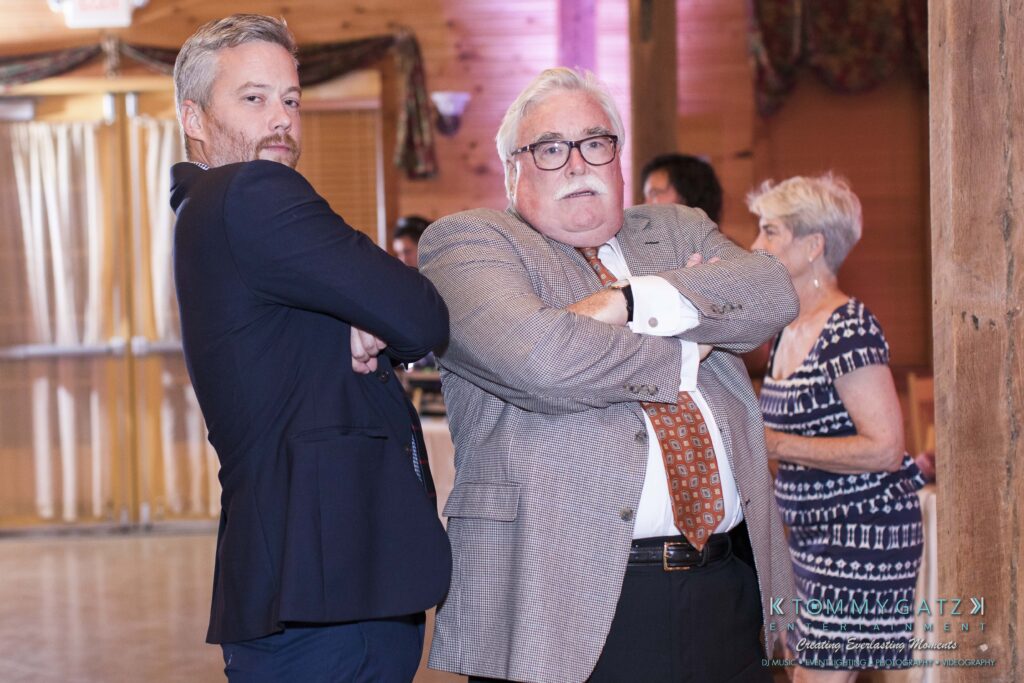 JenniferRudiWeddingReceptionPhotos-370 - TOMMY GATZ ENTERTAINMENT Two men in suits stand back-to-back with arms crossed; one has gray hair and glasses. A woman stands in the background.
