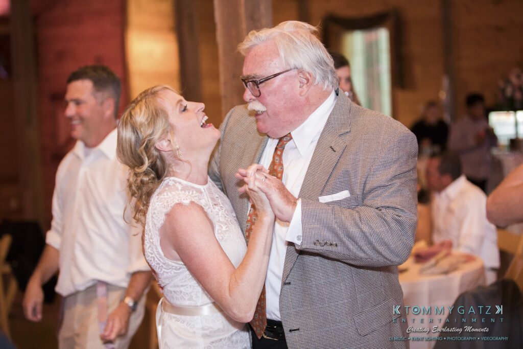 JenniferRudiWeddingReceptionPhotos-375 - TOMMY GATZ ENTERTAINMENT An elderly man and a woman in a white dress joyfully dance together indoors, while another man smiles in the background.