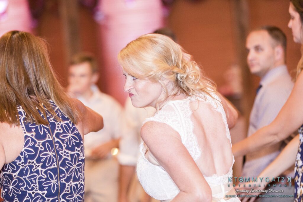 JenniferRudiWeddingReceptionPhotos-376 - TOMMY GATZ ENTERTAINMENT A woman in a white dress dances energetically at a social event, surrounded by other people in a dimly lit room.