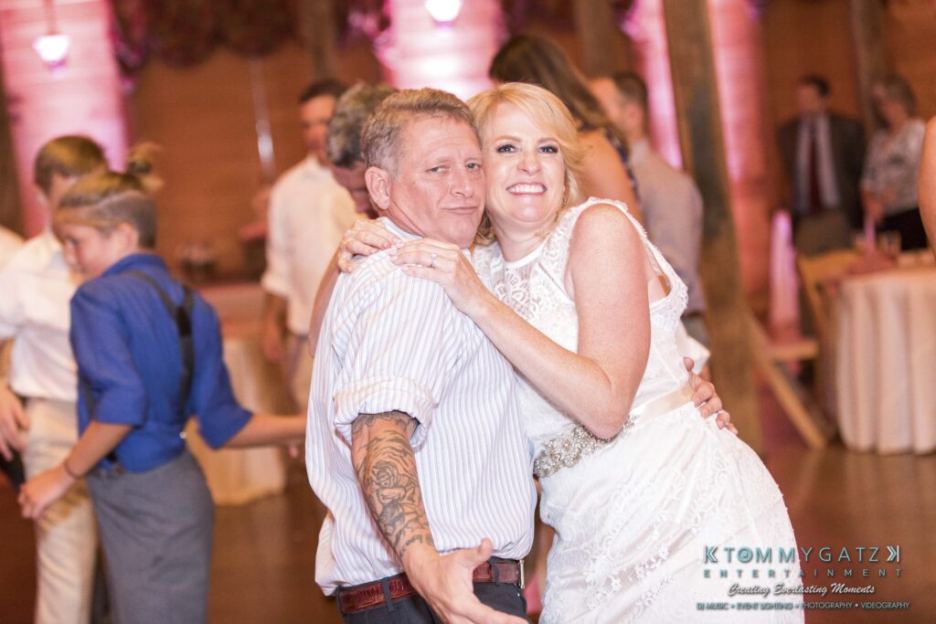 JenniferRudiWeddingReceptionPhotos-377 - TOMMY GATZ ENTERTAINMENT A smiling couple poses closely together in a wedding reception setting. The man has tattoos and wears a striped shirt, while the woman is in a white dress. Guests are visible in the background.