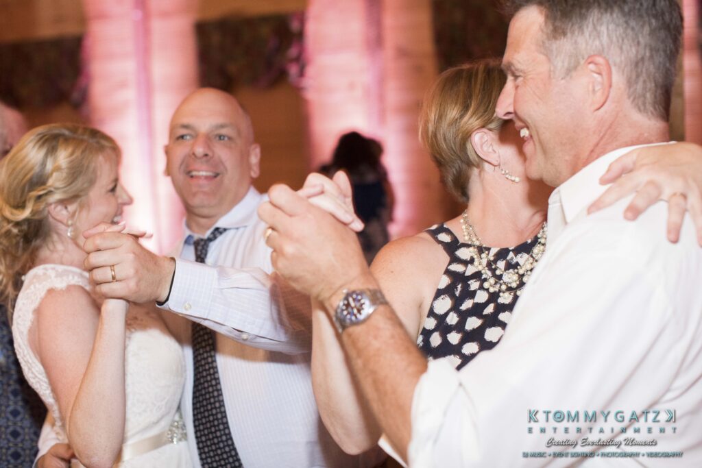 JenniferRudiWeddingReceptionPhotos-382 - TOMMY GATZ ENTERTAINMENT Two couples are dancing at an indoor event. They appear to be smiling and enjoying the moment. The background is softly lit.