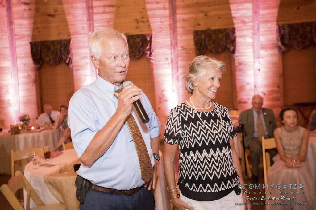 JenniferRudiWeddingReceptionPhotos-383 - TOMMY GATZ ENTERTAINMENT An older man speaks into a microphone while standing next to a smiling older woman in a wooden room set for an event.