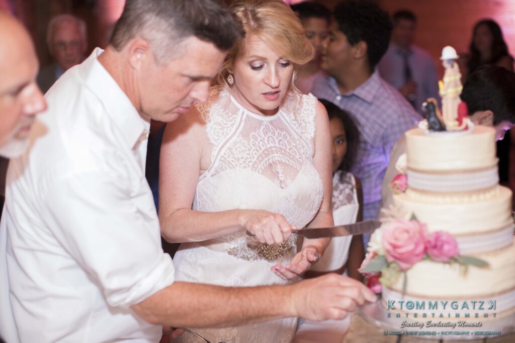 JenniferRudiWeddingReceptionPhotos-386 - TOMMY GATZ ENTERTAINMENT A woman in a white dress cuts a multi-tiered cake with the assistance of a man in a white shirt at a reception.