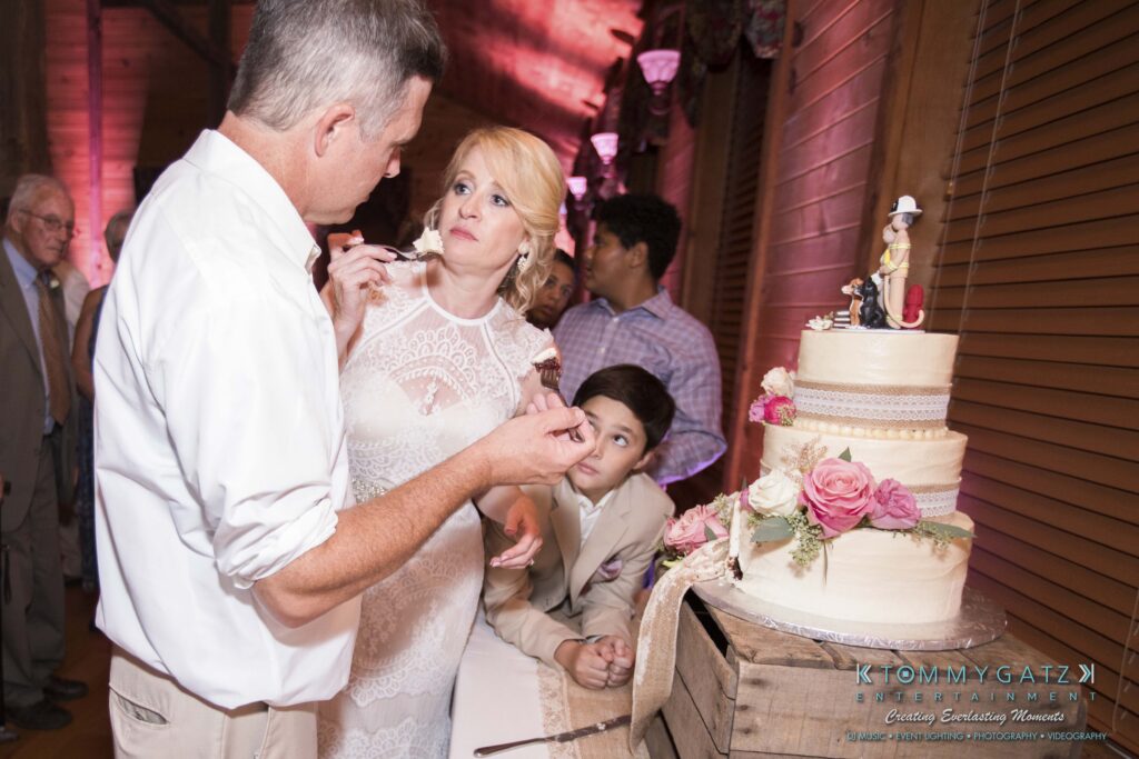 JenniferRudiWeddingReceptionPhotos-388 - TOMMY GATZ ENTERTAINMENT A man and woman stand near a wedding cake with floral decorations. A young boy leans over the table in the background, holding his chin in his hand. Guests are visible in the background.
