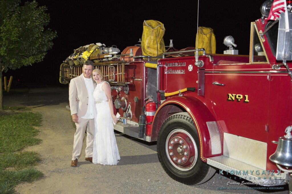 JenniferRudiWeddingReceptionPhotos-399 - TOMMY GATZ ENTERTAINMENT A bride and groom stand smiling next to a vintage red fire truck labeled "No. 1" outdoors at night.