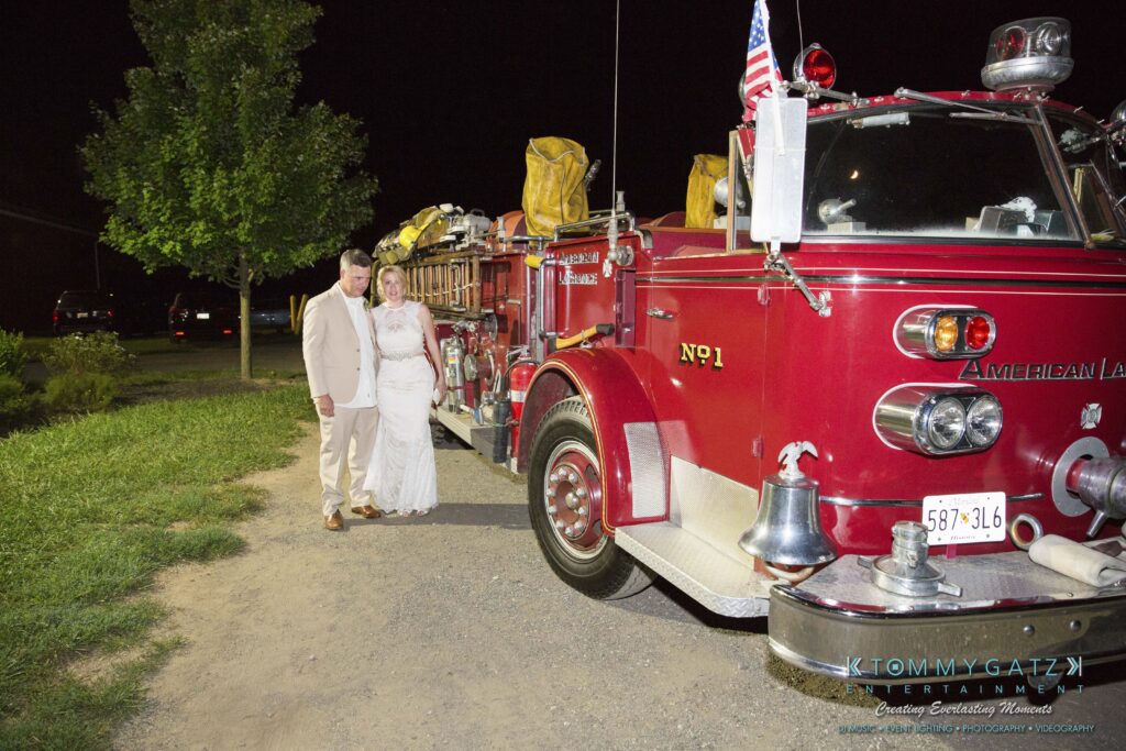 JenniferRudiWeddingReceptionPhotos-400 - TOMMY GATZ ENTERTAINMENT A bride and groom stand smiling beside a vintage red fire truck adorned with American flags, parked on a dirt road at night with a tree and parked cars in the background.