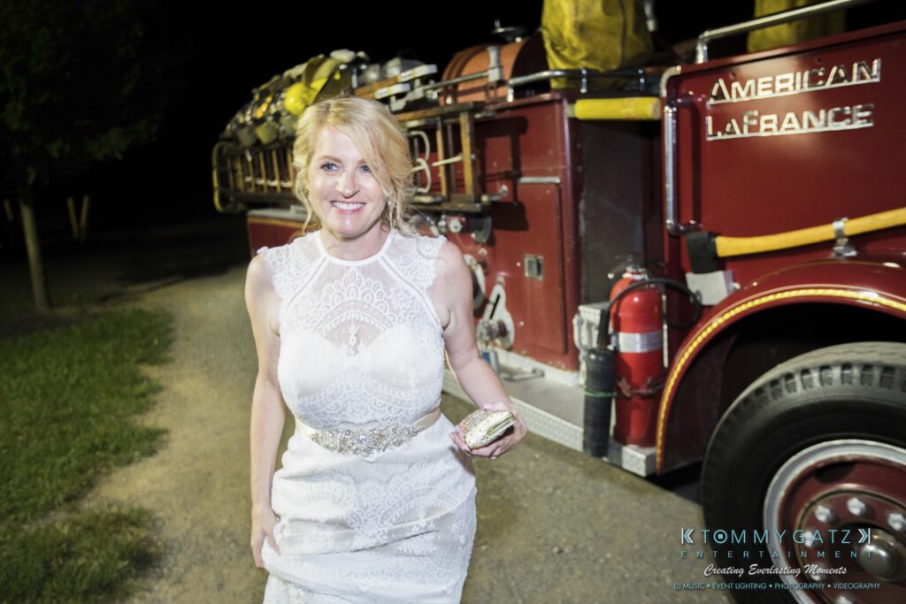 JenniferRudiWeddingReceptionPhotos-404 - TOMMY GATZ ENTERTAINMENT Woman in a white dress standing beside a vintage red fire truck at night, holding a small clutch.