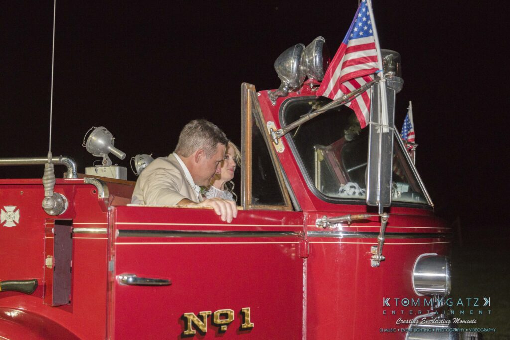 JenniferRudiWeddingReceptionPhotos-405 - TOMMY GATZ ENTERTAINMENT A couple sitting in a vintage red fire truck decorated with U.S. flags at night.