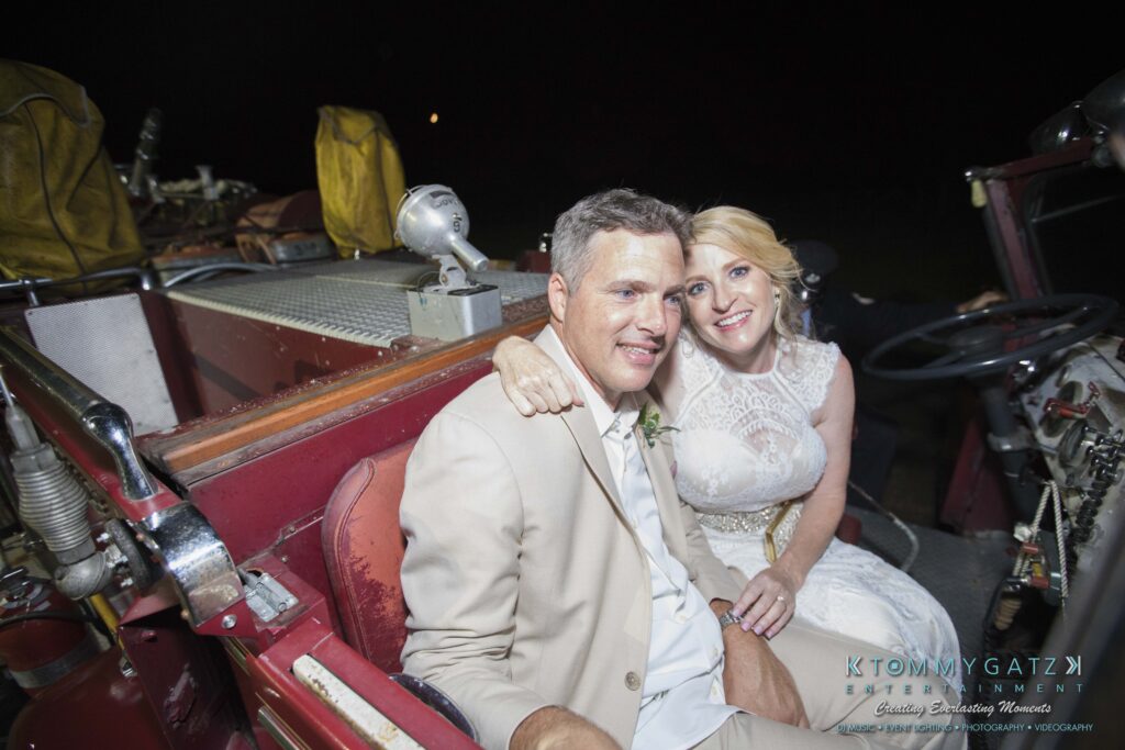 JenniferRudiWeddingReceptionPhotos-407 - TOMMY GATZ ENTERTAINMENT A couple in wedding attire sits closely in an old-fashioned, red car at night.