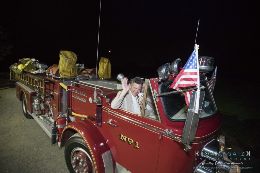 JenniferRudiWeddingReceptionPhotos-409 - TOMMY GATZ ENTERTAINMENT A man waves from the driver's seat of a vintage red fire truck at night. The truck has "No. 1" painted on it and displays an American flag.