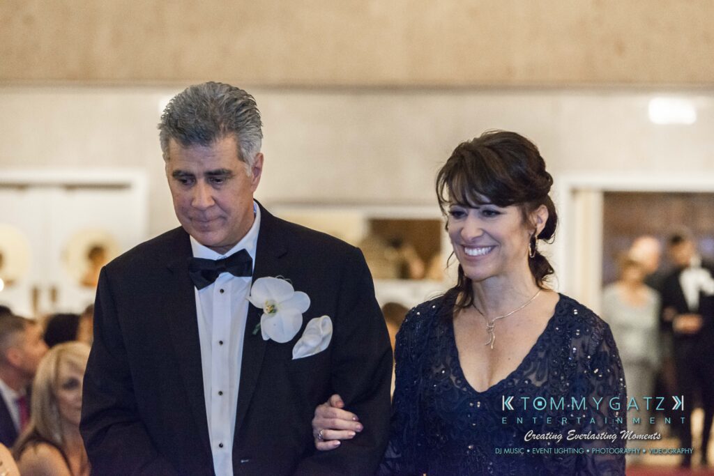 A man in a tuxedo and woman in an evening gown walk arm in arm at a formal event, with others seated in the background.