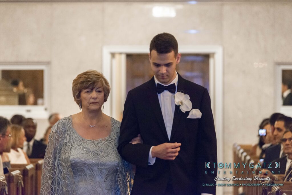 A man in a black tuxedo escorts an older woman in a light blue dress down an aisle.