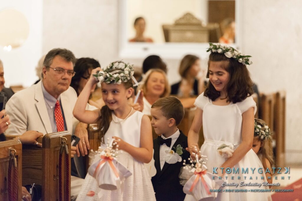Children dressed formally stand in a church holding flower baskets, with seated guests watching them.