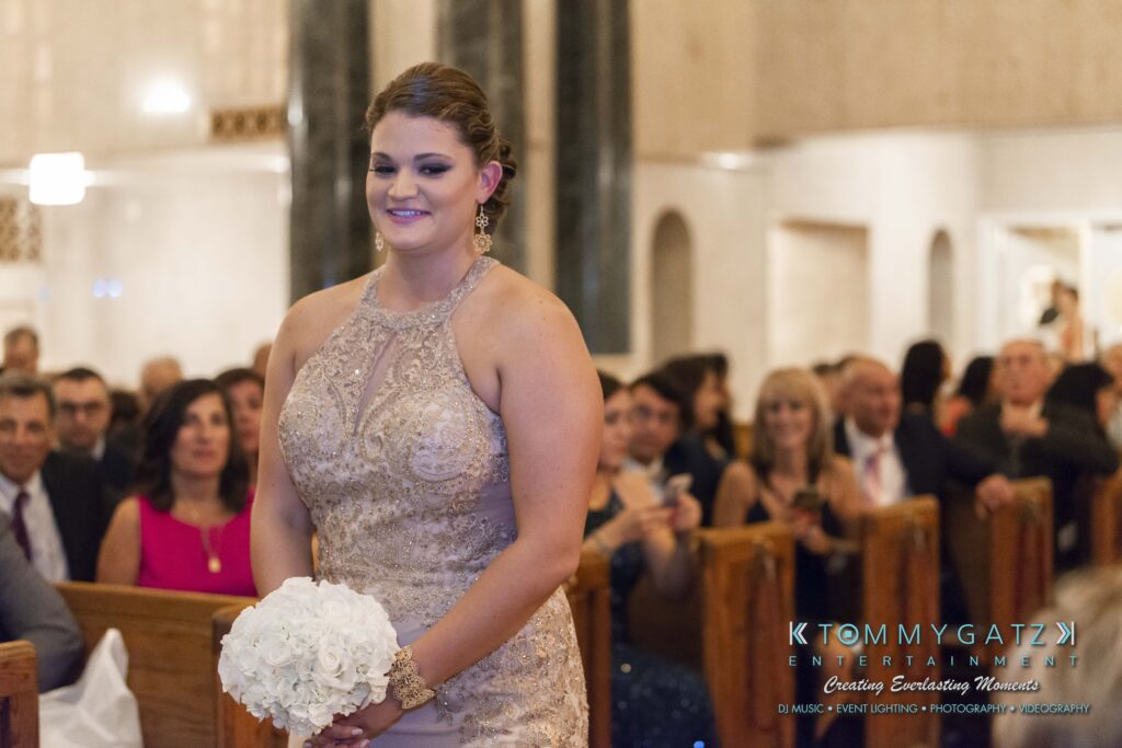 A woman in a sleeveless gown holds a bouquet, walking indoors with seated guests in the background.