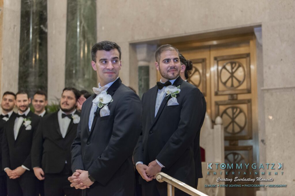 Two groomsmen in tuxedos with white boutonnieres stand in front of a decorative doorway. Other people in formal attire are visible in the background.