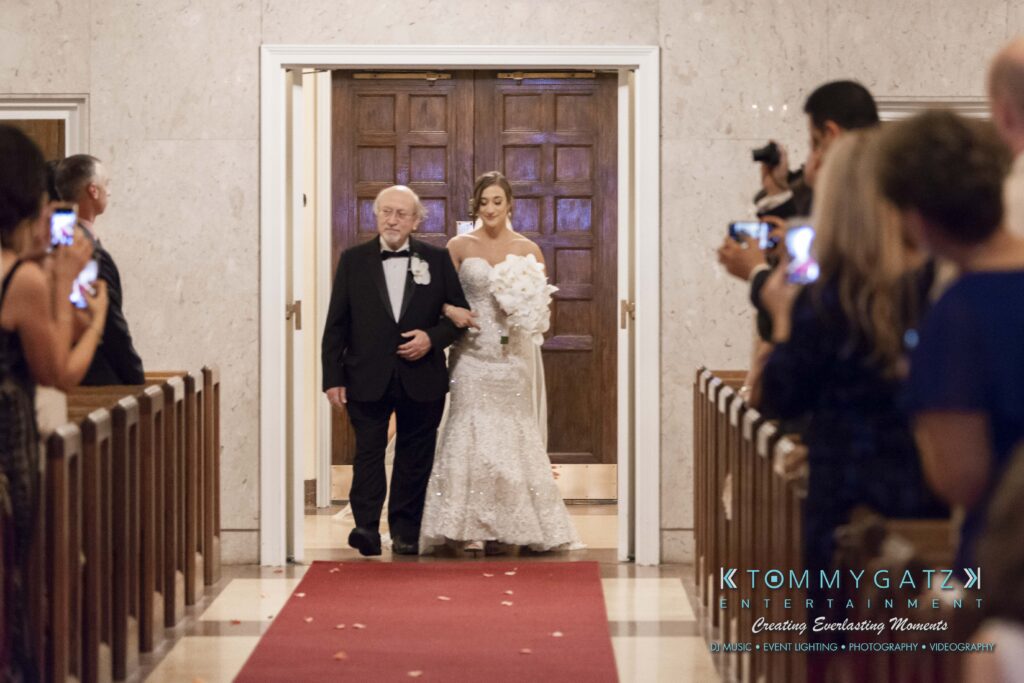 A bride in a white dress walks down the aisle with an older man in a tuxedo, as guests watch and take photos.
