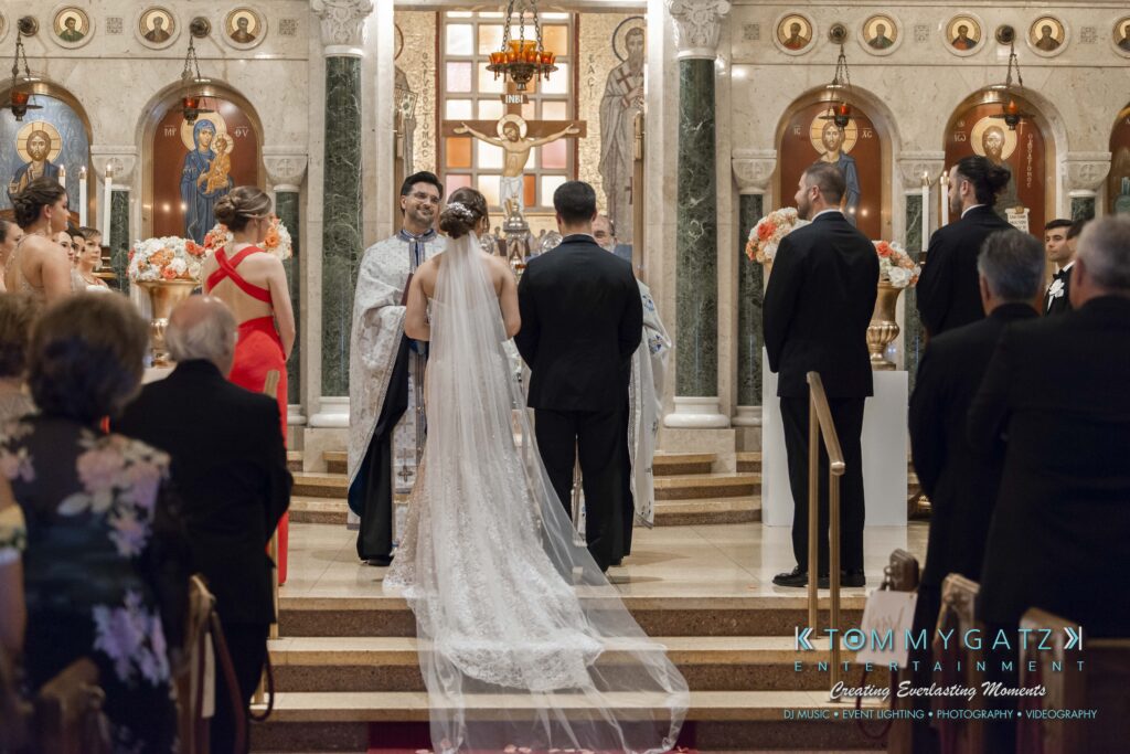 A wedding ceremony inside a church, with a couple standing at the altar, surrounded by attendees. The officiant is speaking, and the church is decorated with icons and flowers.