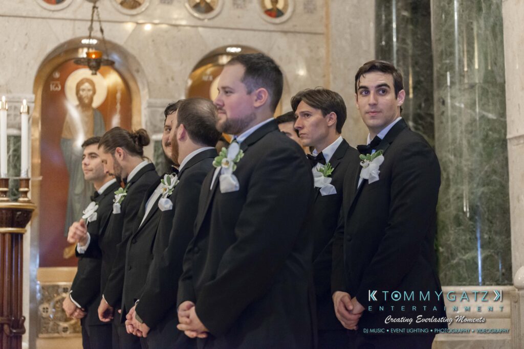 A line of groomsmen in black suits and white boutonnieres stand inside a church, facing forward. Marble walls and religious icons are visible in the background.