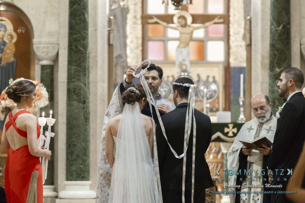 Bride and groom stand before a priest during a wedding ceremony, with crowns being placed on their heads. Two attendants stand nearby.