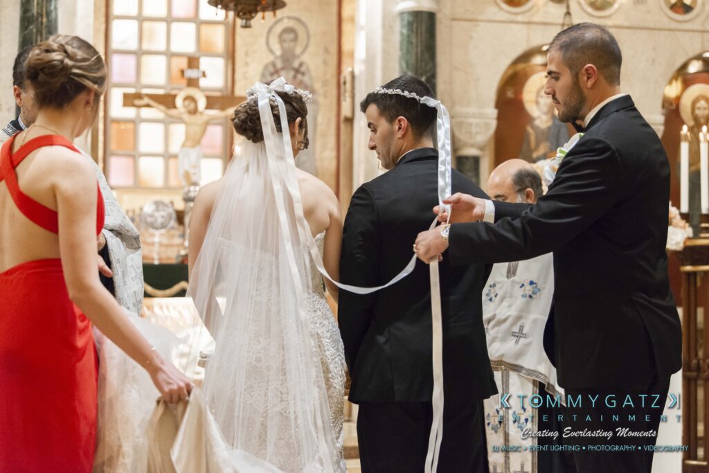A wedding ceremony taking place indoors, with a bride and groom facing forward. A priest is wrapping ceremonial ribbons around their heads, and bridesmaids are standing nearby.