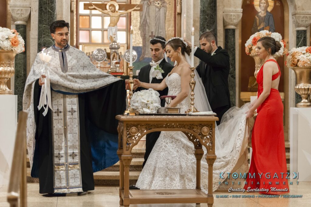 A wedding ceremony in a church with a priest, a bride in a white gown, and a groom in a tuxedo. Two attendants assist the couple, and a woman in a red dress stands nearby.