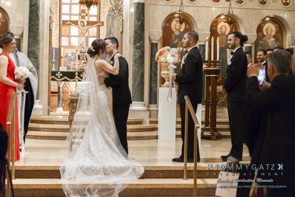 A bride and groom kiss at the altar during a wedding ceremony in a church, with bridesmaids and groomsmen looking on.