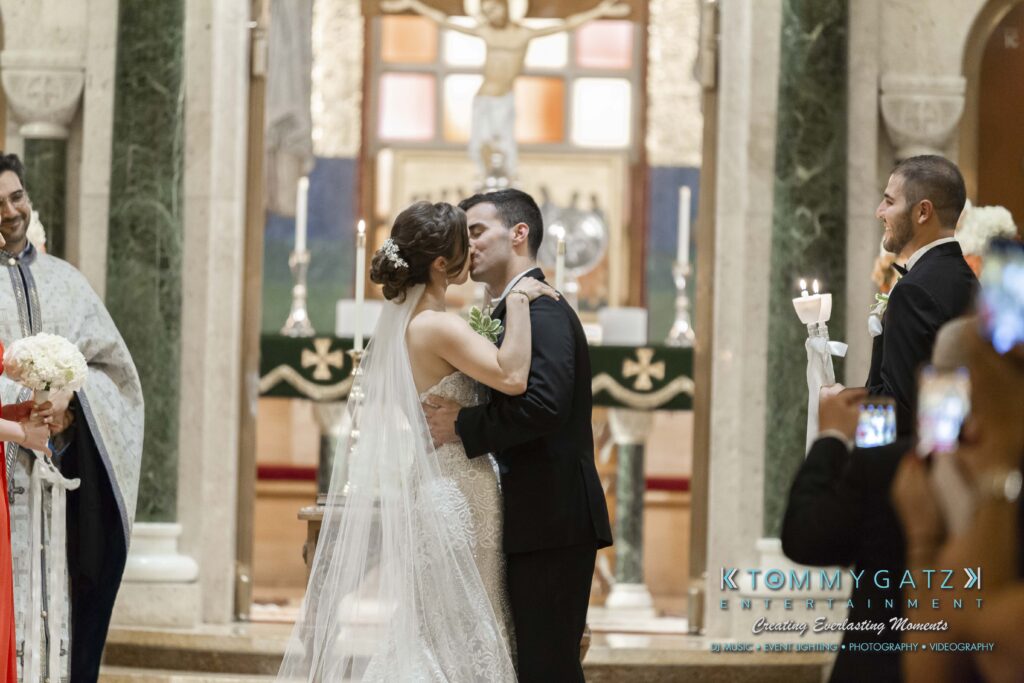Bride and groom share a kiss in a church during their wedding ceremony, surrounded by a priest and attendants.