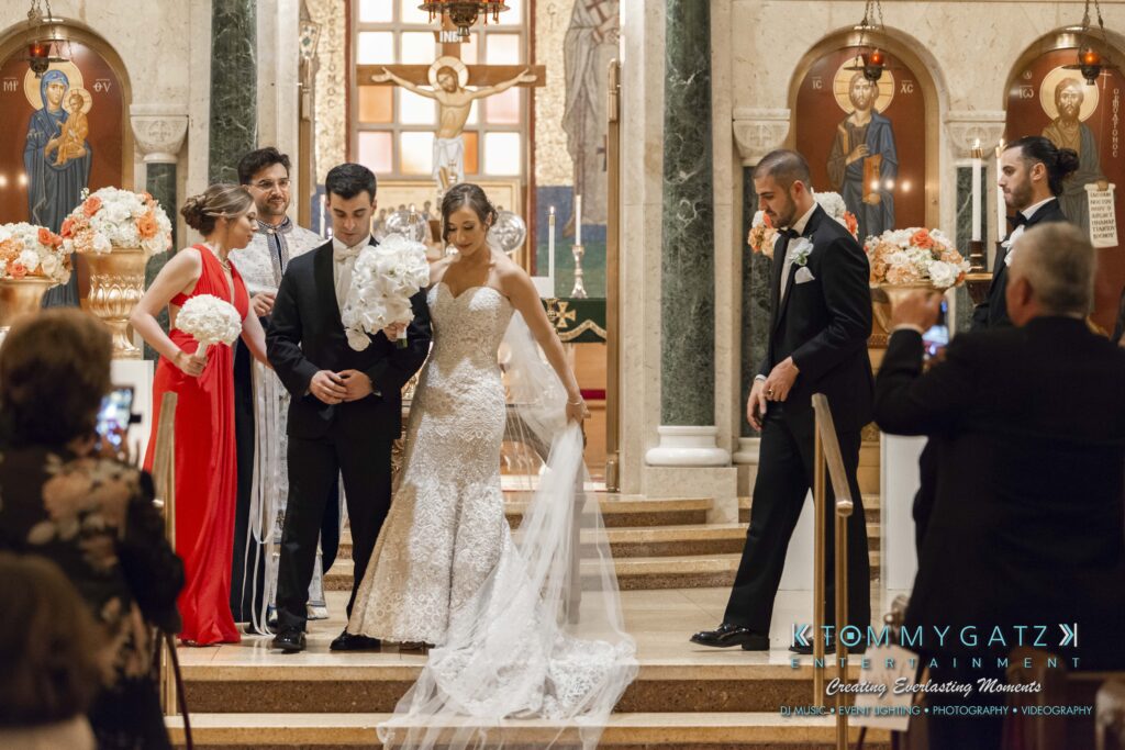 A bride and groom stand together in a church, surrounded by their wedding party. The groom holds a bouquet, and a woman in red stands nearby. Floral arrangements and religious icons decorate the space.