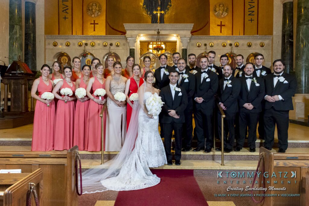 Bride and groom stand with a large wedding party at the altar of a church. Groomsmen in black suits and bridesmaids in pink dresses hold white bouquets.