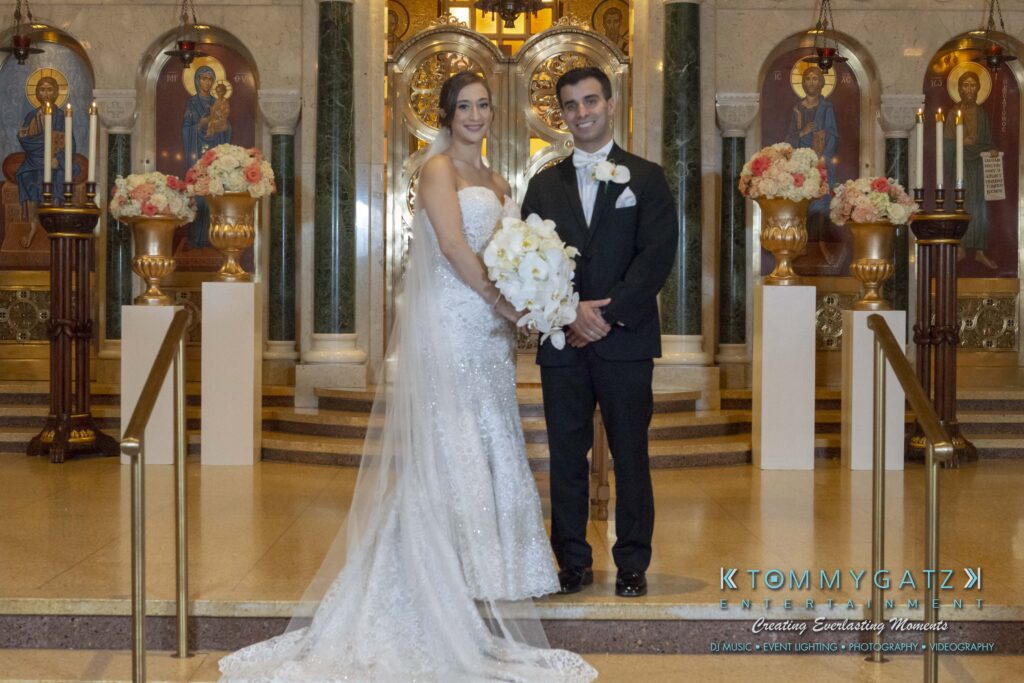 Bride and groom standing together in a church, both smiling and holding a bouquet. They are dressed in formal wedding attire.