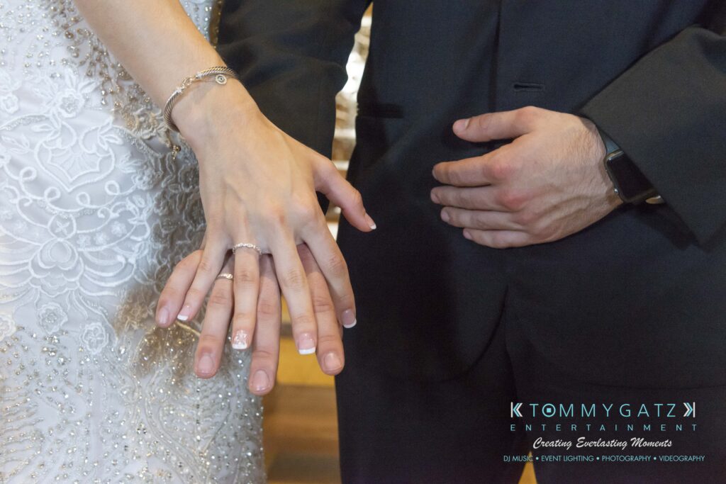 A bride and groom display their wedding rings; the bride wears a white dress, and the groom is in a black suit.