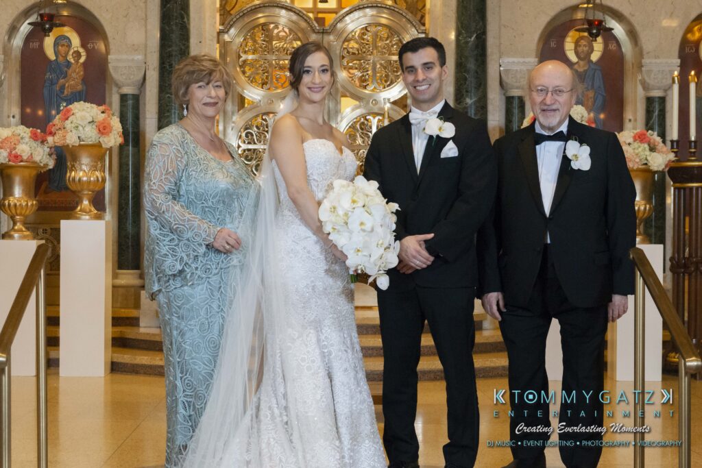 A wedding couple stands in a church with two older adults. The bride holds white flowers, and all are dressed in formal attire.