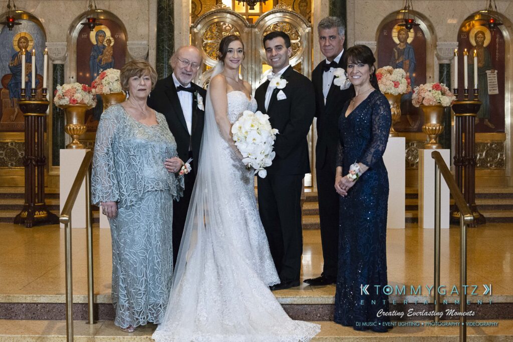 Bride and groom standing with four other people in formal attire inside a decorated venue with ornate background.