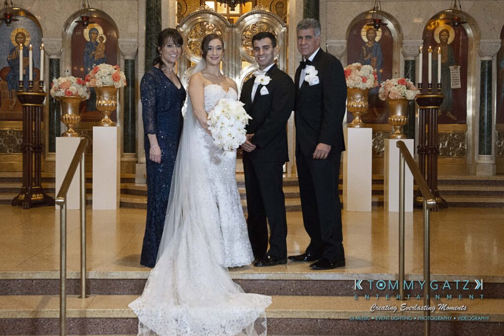 A bride and groom stand with two older adults in formal attire, posing inside a decorative church with floral arrangements.