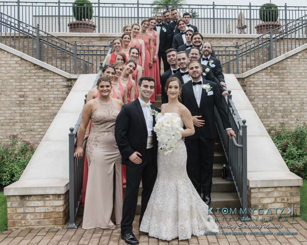 Urania-Aristidis-060218-FORMALS-Low-Res-WM-555 - TOMMY GATZ ENTERTAINMENT A large wedding party posing on outdoor steps. The bride is in a white gown holding flowers, and the groom in a black tuxedo stands beside her. Bridesmaids and groomsmen are arranged in pairs.
