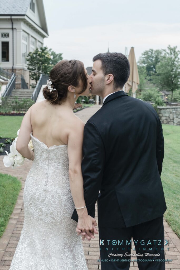 A bride and groom in wedding attire kiss while holding hands, walking on a brick path with a house and garden in the background.