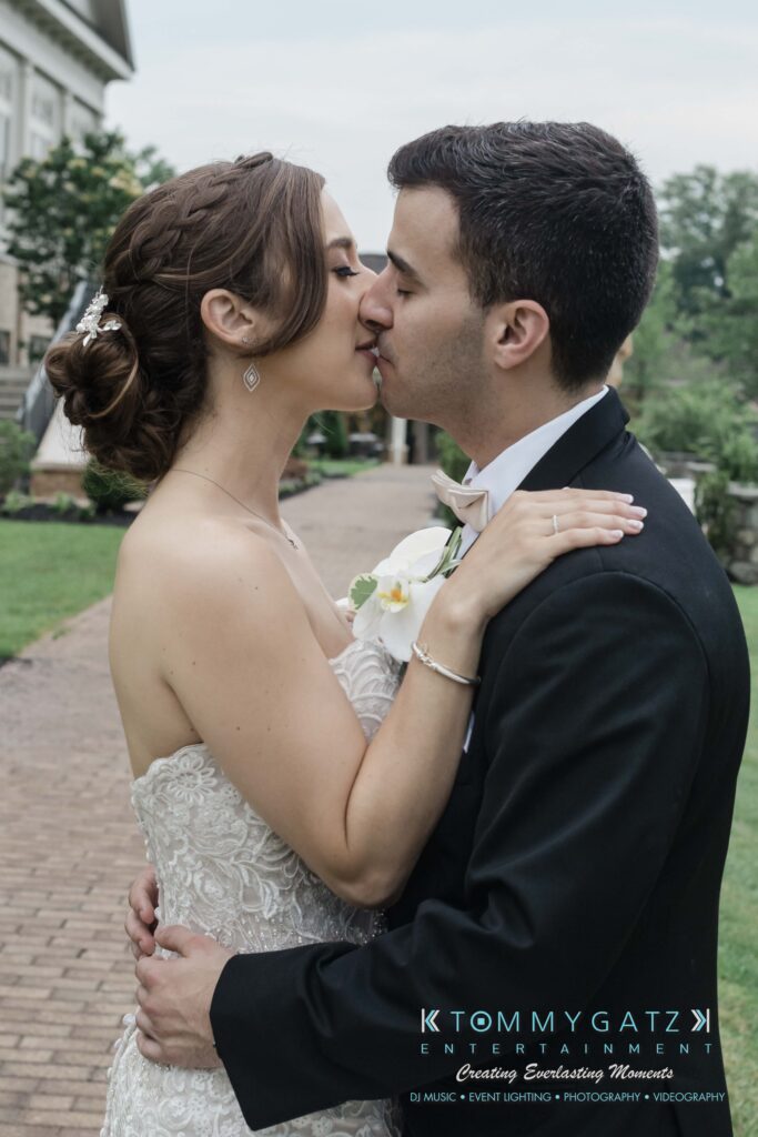 A couple in formal attire shares a kiss outdoors on a pathway, surrounded by greenery.