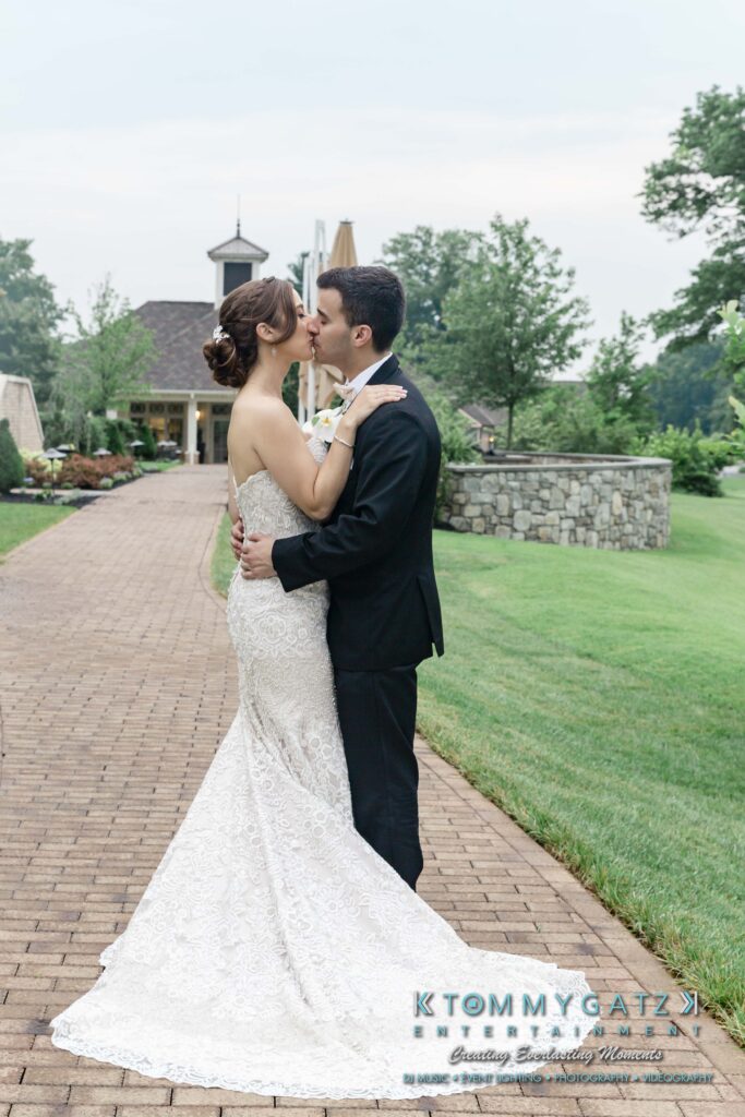 A bridal couple in formal attire shares a kiss outdoors on a walkway, with a building and trees in the background.