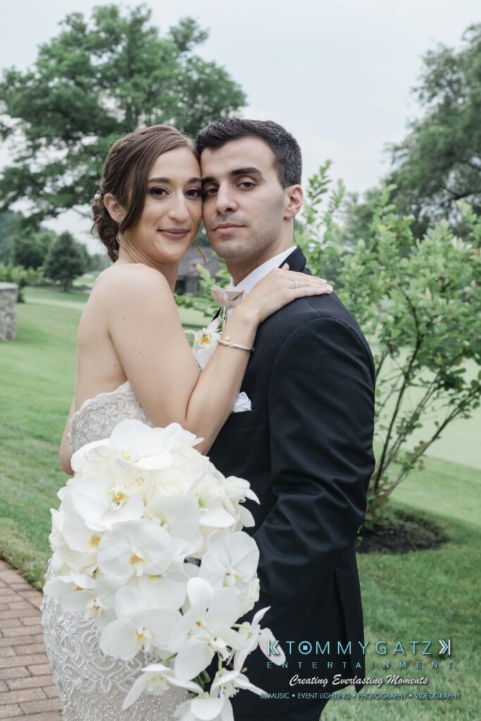 A bride and groom embrace outdoors, with greenery in the background. The bride holds a bouquet of white flowers.