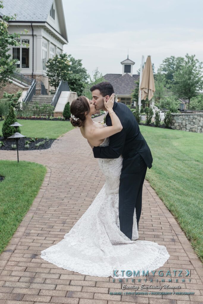 A couple in wedding attire share a dance on a brick path in a garden setting, with houses and greenery in the background.